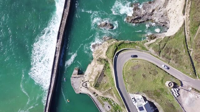 Cars Driving In Lighthouse Hill By Curve Road With Ocean Waves On Pier Near Village Of Portreath, Cornwall, England In Summer. - aerial top down