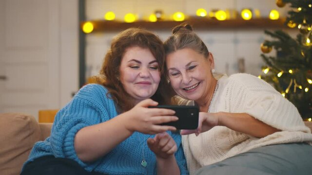 Aged woman and adult daughter resting on couch and watching photo on cellphone on christmas holidays