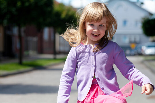 Happy Little Child Running Towards The Camera Jumping. Smiling Cheerful Active Girl With Dispelled Flying Hair Moving Fast Towards The Camera, Closeup, Portrait, Small City In The Back, Outdoors Scene