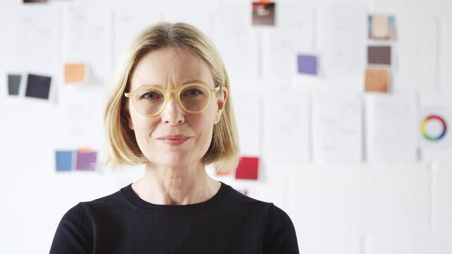 Portrait of smiling mature female fashion designer wearing glasses standing in front of wall in studio next to drawings and swatches - shot in slow motion