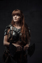 Fototapeta na wymiar Beautiful but dangerous warlike woman viking with long brown hairs in dark armour holding huge axe in dark background.