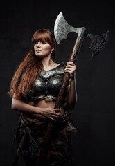 Obraz na płótnie Canvas Dressed in dark armour female scandinavian barbarian with brown hairs holding two handed axe in dark background.