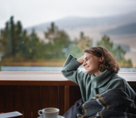 Young happy woman in green sweater by the window in a country house chalet with a view of the mountains chalet - obrazy, fototapety, plakaty