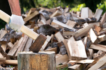 Male hand holds a chopper axe stuck in a stump with a pile of chopped firewood on the background outdoors in summer