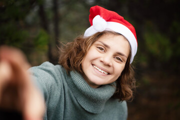 Beautiful happy young woman in santa claus hat in pine forest taking selfie christmas and new year portrait
