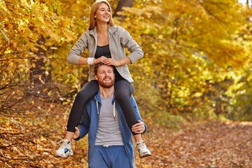 Fototapeta premium cheerful couple spend the day in the autumn forest, blonde female sits on male's neck and smile, have fun, smile looking away