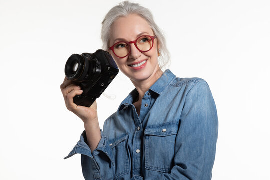Smiling stylish energetic mature female photographer in glasses posing with digital camera on white background. Portrait of positive gray haired middle aged lady. Free time, age and hobby concept.