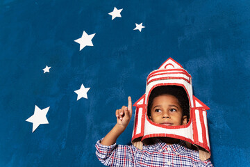 Cardboard rocket helmet on afro-american kid boy and stars on blue background. Clever child with hand made spacecraft playing in cosmonaut. Future astronaut pointing up by index finger.