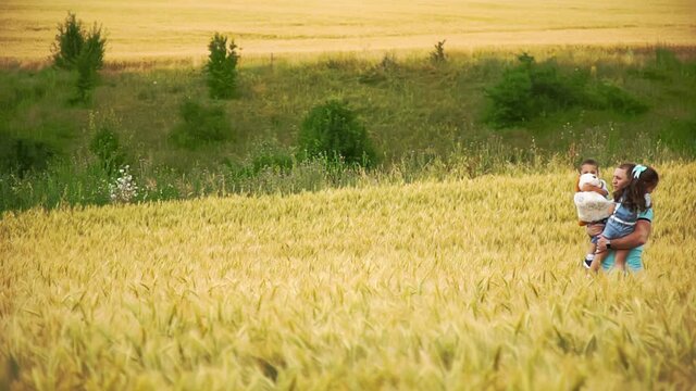 The family walks through the summer field in search of picnic place in slow motion. Father carries two little children on his arms. Pregnant mother is going right behind them with pannier and blanket.