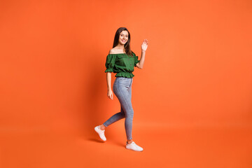 Full length photo of adorable sweet lady wear green off-shoulders blouse walking waving arm isolated orange color background