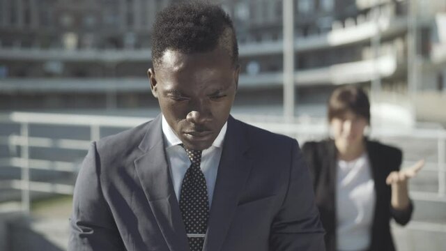 Portrait of upset African American man standing outdoors and thinking as blurred Caucasian woman yelling and gesturing at the background. Sad young businessman having relationship problems.