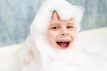 The boy smiles in the soapy foam and washes in the bath. Hygiene, cleanliness concept.