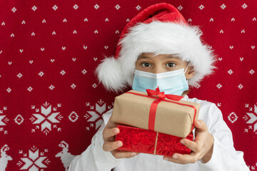 cute little boy in protective face mask and santa hat hold wrapped giftboxes on red christmass background, new reality, holidays in pandemic, greetings concept