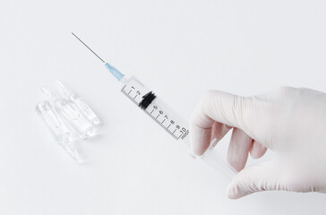 A hand in a white medical glove holds a syringe. In the background, medicine in vials.