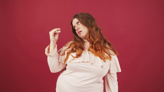 Annoyed oversize lady showing bla bla bla gesture and grimacing, looking at hand with irritation, red background