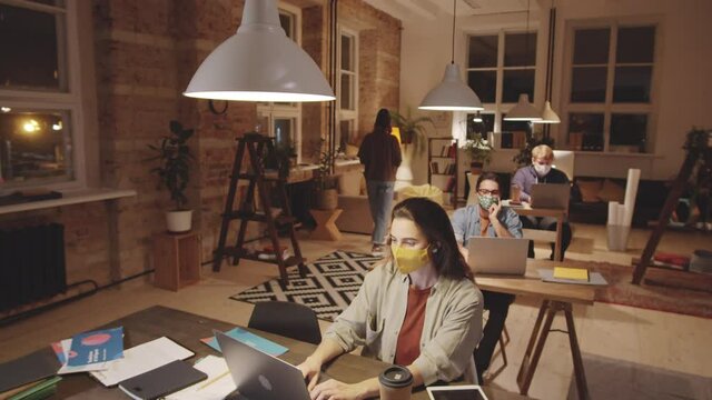 Tilt down shot of young male and female employees in casualwear and protective face masks working in open space loft office in the evening during coronavirus outbreak