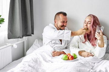 Obraz na płótnie Canvas cheerful couple have breakfast on bed at weekend, after waking up. they lie together on bed in bathrobes, eat healthy food