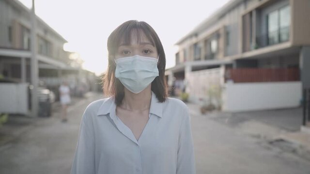 Asian Woman Wear Face Mask Walking In Front Of House Neighborhood Street, Corona Virus Covid19 Awareness Caution, Standing Outdoor Sunset Social Distancing Concept, Showing Confidence By Thumbs Up