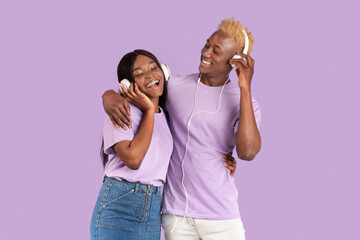 Happy black couple in headphones listening to music and hugging over lilac studio background