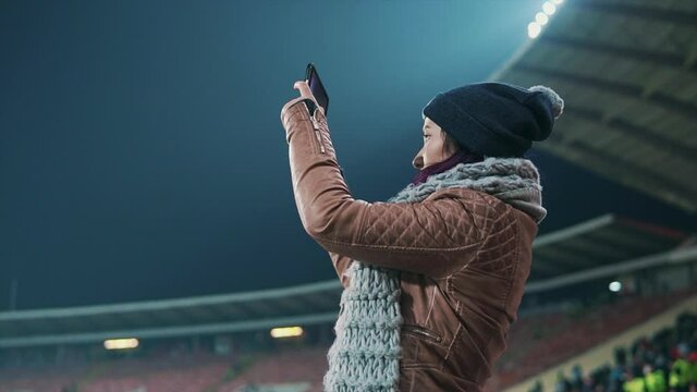 Girl fan taking picture of football match. Female spectator watching sport game at open air illuminated with stadium light. Entertainment, leisure activity and holiday concept.