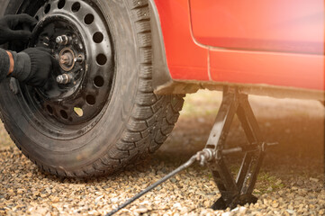 A man twists or unscrews a wheel with a studded tire from a red car standing on a Jack. The concept of puncturing a tire or replacing a summer wheel with a winter one with your own hands