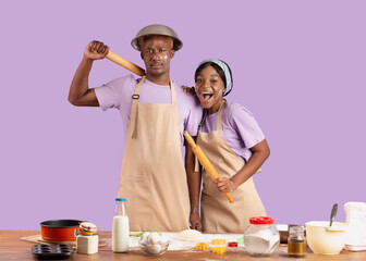 Playful black couple with cheeks covered in flour holding rolling pins, having fun while baking, lilac background