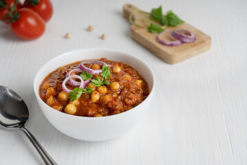 Chole masala or chana indian vegan street food made of chickpeas, tomatoes and cumin decorated with onion rings and parsley served in bowl with spoon on white wooden background. Image with copy space
