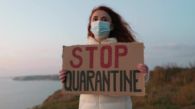 Woman protest holds hands cardboard Stop Quarantine with sign in covid-19 medical mask against blue sunset sky, sea in evening in winter slow motion. Quarantine. Lockdown. Coronavirus. Epidemic