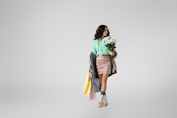 smiling young woman in stylish winter outfit posing with flowers and shopping bags on grey background