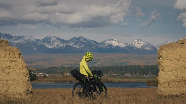 The woman travel on mixed terrain cycle touring with bikepacking. The traveler journey with bicycle bags. Sport bikepacking, bike, sportswear in green black colors. Mountain snow capped, stone arch.