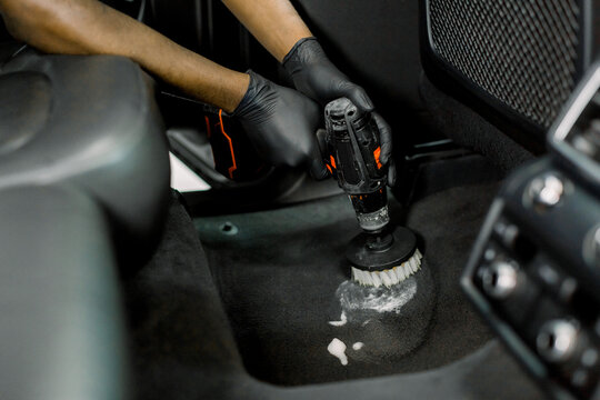 Cropped Close Up Image Of Male Hands, Worker Of Auto Service Station, Cleaning Car Floor And Interior With Disinfecting Foam And Electric Brush Cleaner. Car Detailing Concept