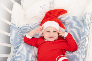 lovely toddler boy in Santa hat, red suit smiling in cot at home.