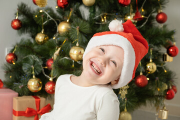 close up portrait little girl in Santa hat laughs near Christmas tree.