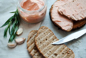 Salmon pasta in a glass jar, served with chopped fried bread, knife, garlic and herbs, black pepper, sea salt on a linen linen napkin on a gray background texture.