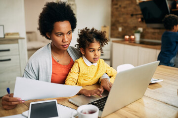 Black working mother using laptop while being with her daughter at home.