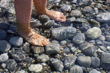 Male bare feet in water on underwater stones in soft focus due to light ripples on water on sunny day