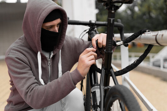 Caucasian male Thief in balaclava and hood breaks the lock on a bicycle in the street during the day. Closed face and hacking. Stealing bicycles.