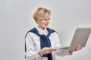 Portrait of elegant middle aged caucasian woman wearing business attire holding and using laptop, standing isolated over grey background