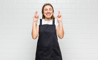 blonde woman smiling and anxiously crossing both fingers, feeling worried and wishing or hoping for...