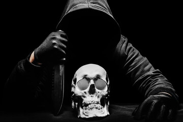 Horror photo of a scary man in hoodie sitting with knife and human skull.