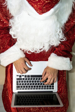 A Young Adult Caucasian Man In A Santa Claus Costume Works At A Laptop, Sitting On The Sofa, Putting The Laptop On His Lap. Top View. Vertical. Close Up. New Year. Christmas. Networking. Cyberspace.