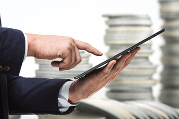 Businessman holds digital tablet and clicks on screen on the background of stacks of coins, close up. Research concept