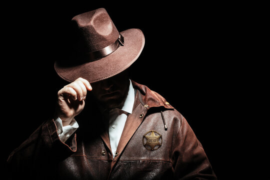 Photo of a shaded sheriff officer with badge in jacket putting on cowboy hat on black background.