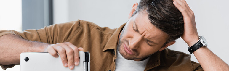 Fototapeta premium Worried man with hand on head looking at broken laptop on blurred background, banner