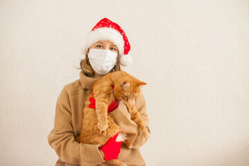 girl in a santa hat and a medical protective mask holds a fat ginger cat in her hands. New Year 2021