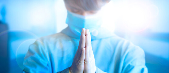 Young female doctor praying with hands clasped together gesture, wearing protective medical face mask and blue uniform. Face close-up. Wide banner with copy space. Health, medicine and pan. 