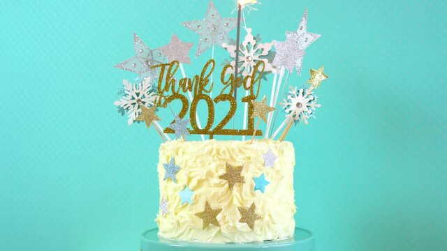 Happy New Year's Eve celebration cake on cake stand in blue white and gold theme decorated with stars and humorous, Thank God It's 2021, cake topper decoration with burning sparkler. Closeup rotating.
