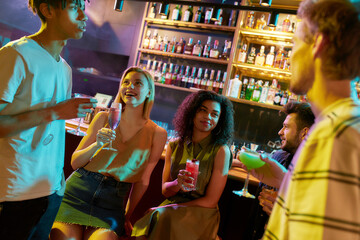 Night Out. Attractive young women drinking cocktail at the bar counter and chatting with men they just met. Young adults having drinks while flirting at the night club