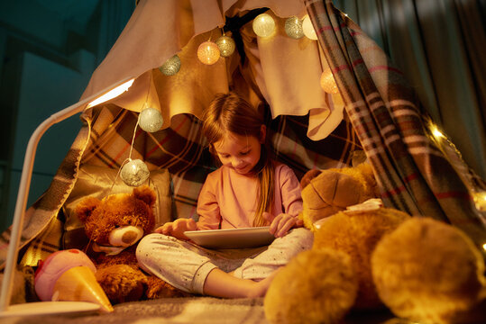 A cute little girl wearing pyjamas sitting on a floor cross-legged barefoot in a self-made hut made of a plaid playing games on a tablet with teddybears and garlands around