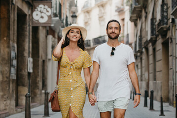 A girl in a yellow dress with a plunging neckline and her boyfriend with a beard are walking in old Spain town. A couple of tourists on a date in the evening Valencia. A lady is checking her hat.